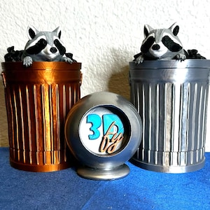 May include: A bronze-colored trash can and a silver trash can, each with a raccoon figurine peering over the top. A silver sphere with the text "3D by" is in the center. The items are on a blue surface.