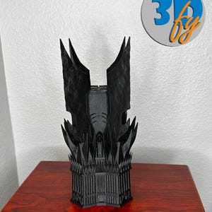 May include: A black 3D-printed model of a fortress with tall spires and intricate architectural details. The model is placed on a dark wood surface. The logo "3D by" is in the upper right corner.