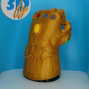 May include: A gold-colored Infinity Gauntlet replica featuring six embedded stones in orange, red, blue, purple, yellow, and green. The gauntlet has intricate mechanical detailing and a circular logo in the top left corner with the text 3D fy.