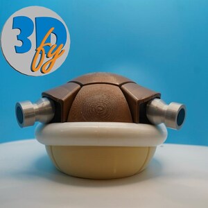 May include: A 3D-printed brown and yellow Koopa shell with two silver-colored cannons. The shell has a white trim and is set against a blue background. The logo "3D by" is in the upper left corner.