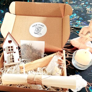 Peut inclure: Une boîte en carton remplie d'objets spirituels : une maison en bois, un parchemin, des herbes, un bâton de palo santo et de petites bouteilles en verre. Un autocollant avec le texte "MAGIC INSIDE" est sur la boîte. Une bougie et une statue de Bouddha sont également présentes.