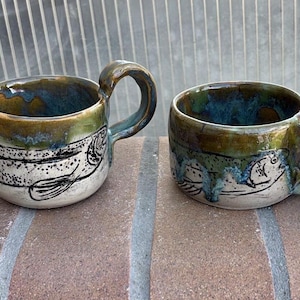 May include: Two ceramic mugs featuring a hand-drawn black fish illustration on a white base. The upper portion of each mug is glazed in a glossy, mottled olive green and blue finish, with a curved handle extending from the side of each vessel.