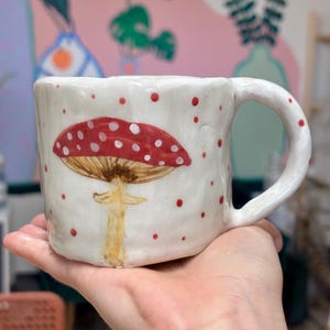 Pode incluir: Uma caneca de cerâmica branca com uma textura irregular moldada à mão. O exterior apresenta um cogumelo vermelho pintado à mão com manchas brancas no chapéu e um caule bege. Pequenos pontos vermelhos estão espalhados pela superfície.