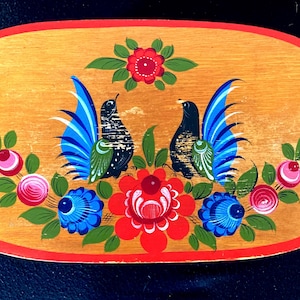 Könnte beinhalten: Ovales Holztablett mit einem bemalten Volkskunstdesign. Zwei schwarze Vögel mit blauen Schwanzfedern blicken sich an, umgeben von roten, blauen und rosa floralen Elementen. Das Tablett hat einen roten Rand und einen hellbraunen Hintergrund.