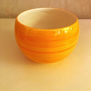 May include: A round ceramic bowl featuring a vibrant orange exterior with subtle horizontal brushstroke patterns in varying shades of yellow and orange. The interior of the bowl is a smooth, solid off-white color, providing a clean contrast to the exterior.