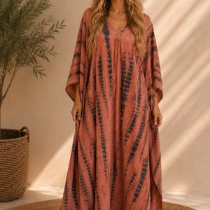 May include: A floor-length kaftan dress in a muted terracotta orange hue featuring a vertical tie-dye pattern in dark charcoal gray. The garment has a V-neckline, wide flowing sleeves, and a loose, relaxed silhouette that drapes to the floor.