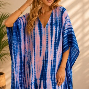 May include: A loose-fitting kaftan dress featuring a vibrant tie-dye pattern with vertical stripes of deep blue and soft light pink. The garment has a deep V-neckline, wide short sleeves, and a relaxed, flowing silhouette that reaches below the knees.