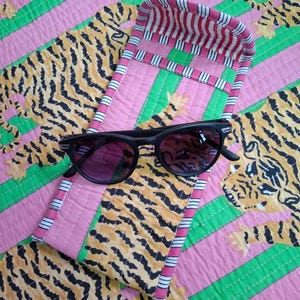 May include: A pair of black sunglasses with dark lenses resting on a quilted fabric case. The case features a repeating pattern of orange and black tigers on a pink and green striped background, with a red and white striped interior lining.