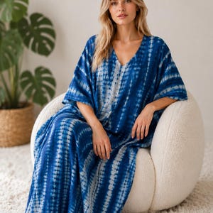 May include: A floor-length kaftan dress featuring a deep blue and white tie-dye pattern with vertical stripes, a V-neckline, and loose, wide sleeves. The garment has a relaxed, flowing silhouette that extends to the floor, showcasing a repeating pattern.
