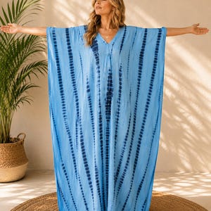 May include: A floor-length kaftan dress in light blue featuring a vertical tie-dye pattern in dark navy blue. The garment has a V-neckline, wide short sleeves, and a loose, flowing silhouette that extends to the ankles with a straight hemline.