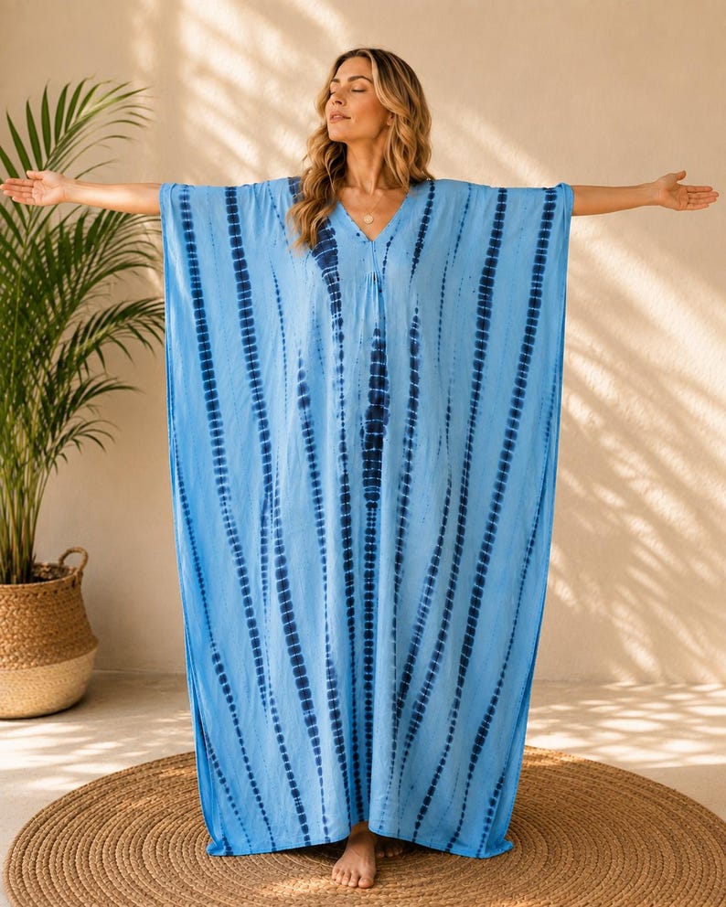 May include: A floor-length kaftan dress in light blue featuring a vertical tie-dye pattern in dark navy blue. The garment has a V-neckline, wide short sleeves, and a loose, flowing silhouette that extends to the ankles with a straight hemline.
