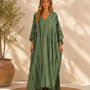May include: A floor-length kaftan dress in olive green featuring a vertical tie-dye pattern in dark charcoal. The garment has a V-neckline, loose-fitting wide sleeves, and side slits at the hem, providing a relaxed and flowing silhouette.