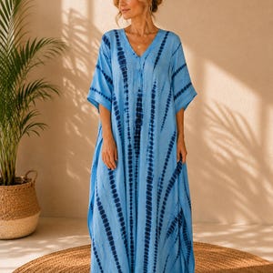 May include: A floor-length kaftan dress in light blue featuring a vertical tie-dye pattern in dark navy blue. The garment has a V-neckline, loose short sleeves, and a relaxed, flowing silhouette that extends to the ankles with side slits.