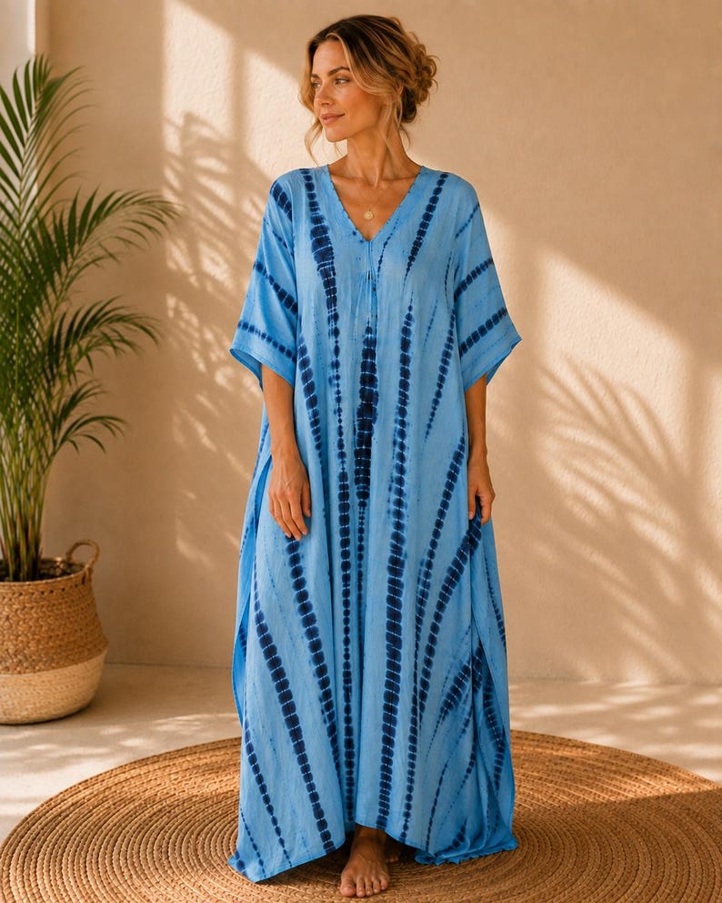May include: A floor-length kaftan dress in light blue featuring a vertical tie-dye pattern in dark navy blue. The garment has a V-neckline, loose short sleeves, and a relaxed, flowing silhouette that extends to the ankles with side slits.