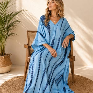 May include: A floor-length kaftan dress in light blue featuring a dark blue tie-dye pattern with vertical stripes. The garment has a V-neckline, loose-fitting short sleeves, and a relaxed, flowing silhouette that reaches down to the floor.