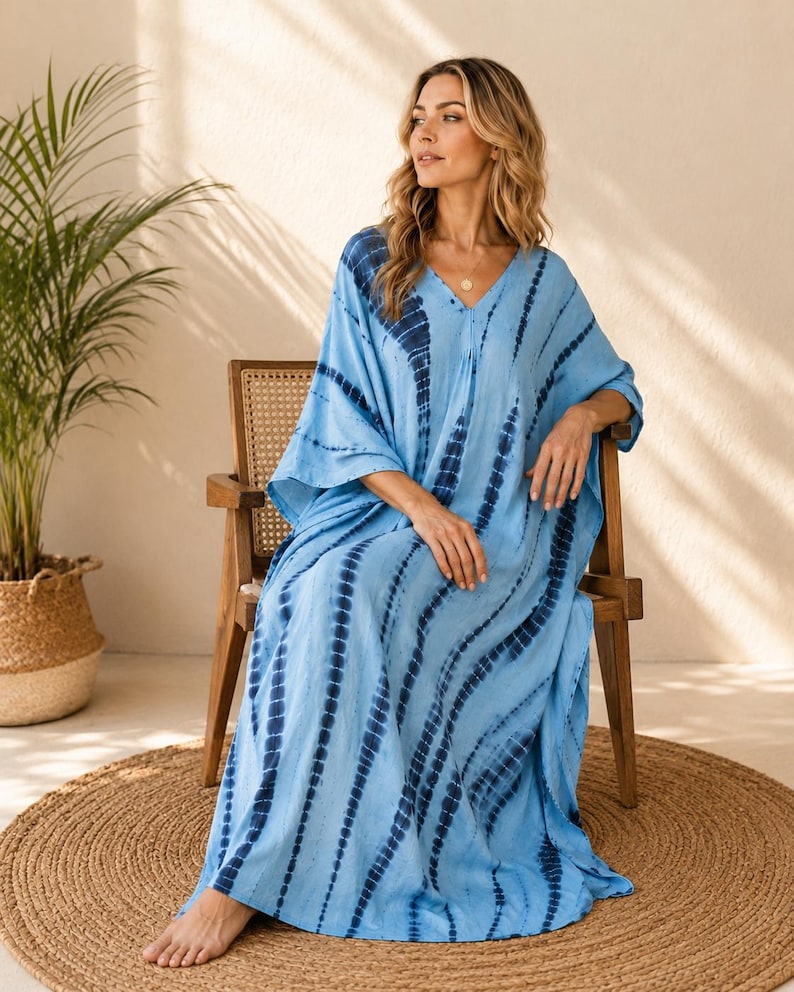 May include: A floor-length kaftan dress in light blue featuring a dark blue tie-dye pattern with vertical stripes. The garment has a V-neckline, loose-fitting short sleeves, and a relaxed, flowing silhouette that reaches down to the floor.