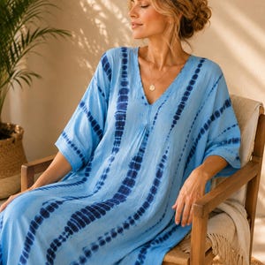 May include: A light blue kaftan dress featuring a vertical tie-dye pattern in dark navy blue. The garment has a V-neckline, loose elbow-length sleeves, and a relaxed, flowing silhouette that extends to the floor, crafted from a lightweight fabric.