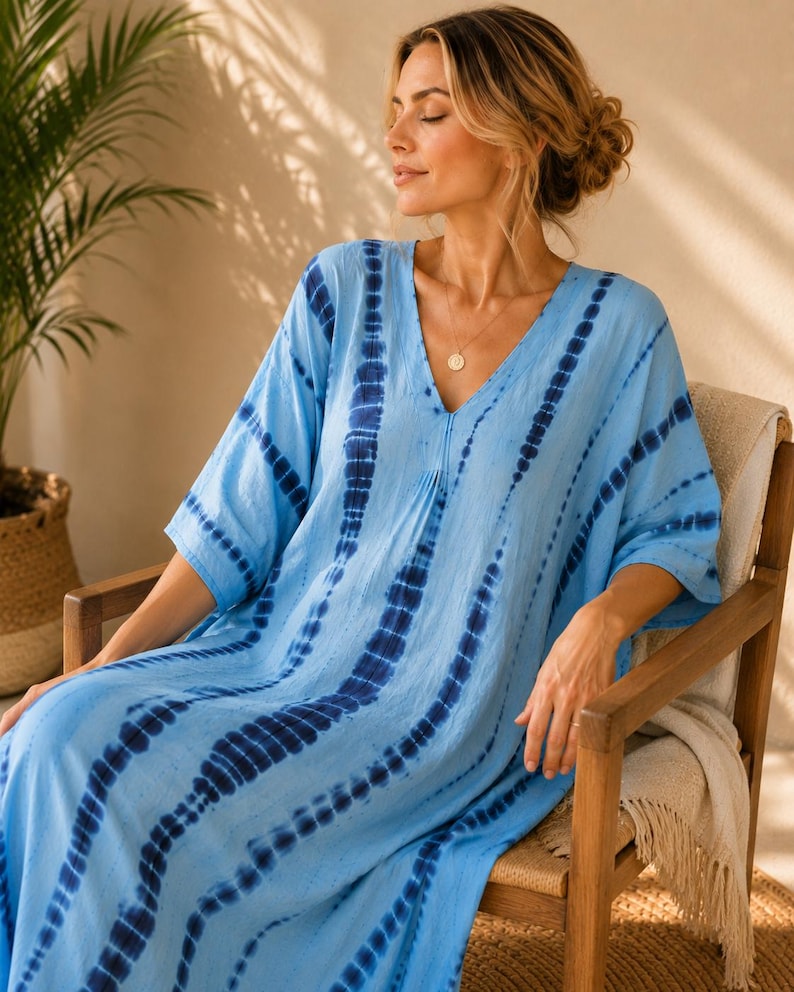 May include: A light blue kaftan dress featuring a vertical tie-dye pattern in dark navy blue. The garment has a V-neckline, loose elbow-length sleeves, and a relaxed, flowing silhouette that extends to the floor, crafted from a lightweight fabric.
