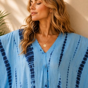 May include: A light blue kaftan featuring a vertical tie-dye pattern in dark navy blue. The garment has a V-neckline and loose, flowing sleeves. A gold-toned circular pendant necklace hangs from the neckline, resting against the fabric.