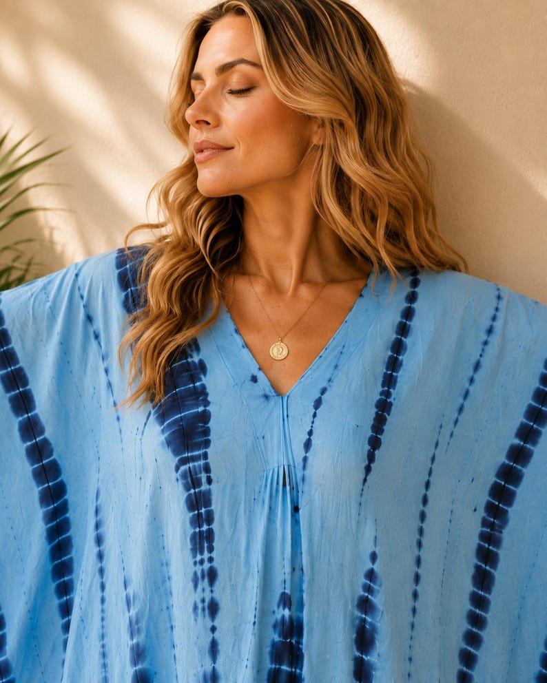 May include: A light blue kaftan featuring a vertical tie-dye pattern in dark navy blue. The garment has a V-neckline and loose, flowing sleeves. A gold-toned circular pendant necklace hangs from the neckline, resting against the fabric.