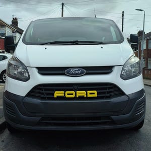 Puede incluir: Una furgoneta Ford Transit blanca con una parrilla y parachoques negros. La palabra "FORD" se muestra en letras amarillas en la parrilla. El logotipo de Ford está centrado encima de la parrilla.