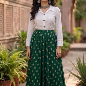 Peut inclure: Une femme porte une chemise blanche boutonnée et une longue jupe verte ornée d'un motif floral doré. Elle est accessoirisée de bijoux argentés. Le fond présente de la verdure et un mur en pierre.
