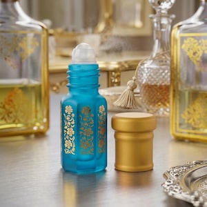 May include: A small, square-shaped, frosted teal glass roll-on bottle featuring gold floral patterns on its sides. The bottle has a clear plastic roller ball top and a separate, solid gold-colored cylindrical cap sitting beside it.