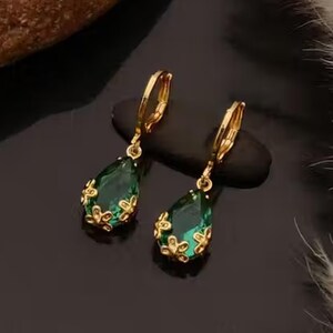 Puede incluir: Un par de pendientes colgantes dorados con piedras en forma de lágrima de color verde esmeralda. Los pendientes tienen un diseño floral alrededor de la piedra verde y un cierre de palanca. Se muestran sobre una superficie negra.