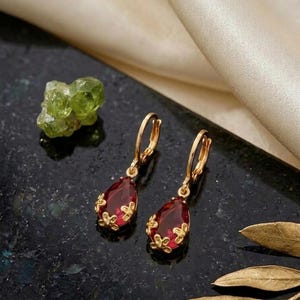 May include: A pair of gold hoop earrings featuring teardrop-shaped red gemstones. Each gemstone is encased in a decorative gold setting with small floral-like metal accents at the base, suspended from a simple gold hoop closure mechanism.