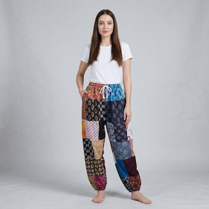 May include: A woman wearing patchwork trousers and a white t-shirt. The trousers are made from various colourful fabric patches with different patterns and colours, including blue, brown, and orange. They have a drawstring waist and pockets.