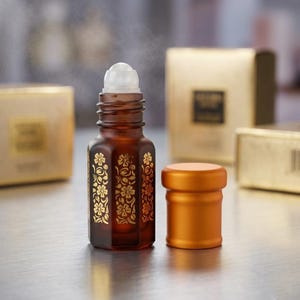 Könnte beinhalten: Eine braune Glas-Roll-on-Flasche mit goldenen Blumenmustern und einem weißen Rollball-Aufsatz. Die Flasche befindet sich neben einer orangefarbenen Kappe. Im Hintergrund befinden sich mehrere goldene Schachteln.
