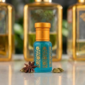May include: A small, rectangular, translucent turquoise glass perfume bottle with a metallic gold cap. The bottle features an intricate gold floral pattern printed on its front surface. A single star anise and two cardamom pods sit nearby.