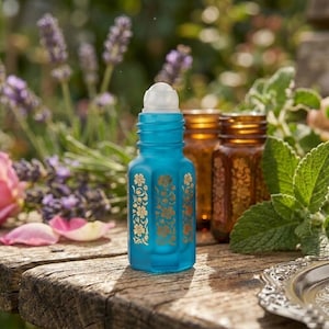 May include: A turquoise glass essential oil roller bottle with a white rollerball top and gold floral designs. Two amber glass bottles are in the background. The bottles are on a weathered wooden surface, with flowers and greenery in the background.
