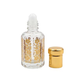 May include: Clear glass roll-on perfume bottle with a white rollerball top and gold floral design. A separate gold-colored cap sits next to the bottle. The bottle has a decorative, vintage aesthetic.