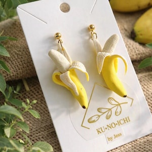 May include: A pair of dangling earrings shaped like peeled yellow bananas with white inner peels, attached to gold-tone ball studs. The earrings are mounted on a white display card featuring a gold triangular logo and the text KU-NO-ICHI by Jess.