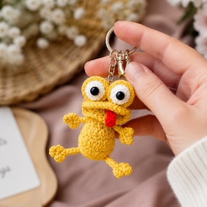 May include: A handmade yellow crochet frog keychain with large googly eyes and a red tongue. The frog is attached to a silver keyring. The background includes flowers and a card with the text "Collect moments not things."