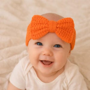 Puede incluir: Primer plano de un bebé sonriente con una diadema de lazo naranja tejida. La diadema está hecha de hilo texturizado y tiene forma de lazo. El bebé lleva un body blanco.