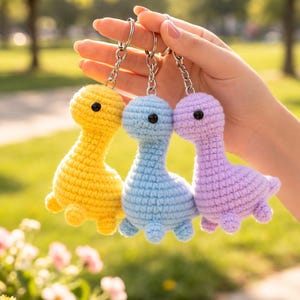 Könnte beinhalten: Drei gehäkelte Dinosaurier-Schlüsselanhänger in Gelb, Blau und Lila. Jeder Dinosaurier hat ein schwarzes Auge und ist an einem silbernen Schlüsselanhänger befestigt. Die Schlüsselanhänger werden vor einem verschwommenen Hintergrund aus grünen Bäumen und Blumen gehalten.