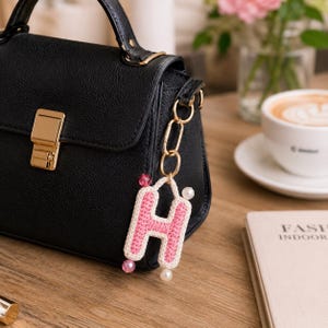 May include: A black leather handbag with a gold clasp and handle. A pink and white crocheted letter "H" charm with pearl and pink bead accents hangs from the bag. The charm is attached to the bag with a gold chain.