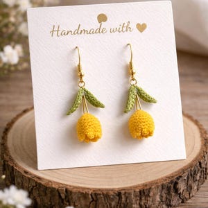 Könnte beinhalten: Ein Paar handgefertigte gelbe Häkeltulpen-Ohrringe mit grünen Blättern und goldfarbenen Haken. Die Ohrringe werden auf einer weißen Karte mit dem Text "Handmade with" und einem Herzsymbol präsentiert.