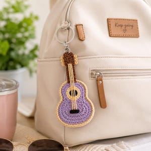May include: A crochet guitar keyring featuring a lavender body with a cream-coloured border, a brown neck, and yellow strings. The keyring is attached to a silver metal ring and is hanging from the zip of a beige backpack with a Keep going label.