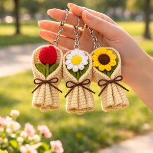 Könnte beinhalten: Drei Schlüsselanhänger mit gehäkelten Blumensträußen. Jeder Schlüsselanhänger zeigt eine andere Blume: eine rote Tulpe, eine weiße Gänseblümchen und eine gelbe Sonnenblume. Die Sträuße sind in beigem Garn gewickelt und mit einem braunen Band gebunden. Die Schlüsselanhänger werden von einer Person gehalten.
