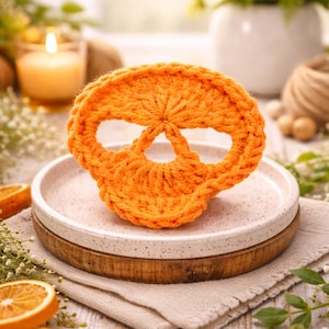 Könnte beinhalten: Ein orangefarbener, gestrickter Totenkopf liegt auf einem gesprenkelten Keramikteller mit Holzsockel. Der Totenkopf hat Augen- und Nasenöffnungen. Der Hintergrund umfasst eine brennende Kerze, Blumen und getrocknete Orangenscheiben.