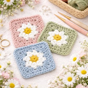 Könnte beinhalten: Drei gehäkelte quadratische Untersetzer in Pastellfarben: Rosa, Grün und Blau. Jeder Untersetzer zeigt eine weiße Gänseblümchenblüte mit gelber Mitte. Das Bild enthält auch Wolle, Häkelnadeln und Blumen, was auf ein handgefertigtes Thema hindeutet.