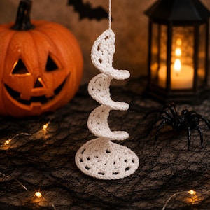 Könnte beinhalten: Ein weißes, gehäkeltes Spiralornament, das aus einer Reihe verbundener kreisförmiger Formen besteht, die sich nach oben winden. Die Struktur ist aus weißem Garn mit einem sichtbaren Lochmuster gefertigt und an einer dünnen weißen Schnur aufgehängt.
