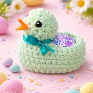 Könnte beinhalten: Ein gehäkeltes Entchen in Hellgrün mit orangefarbenem Schnabel und schwarzen Augen hält ein verziertes Osterei. Ein türkisfarbenes Band ist um seinen Hals gebunden. Pastellfarbene Ostereier und Gänseblümchen umgeben das Entchen auf einer rosa Oberfläche.