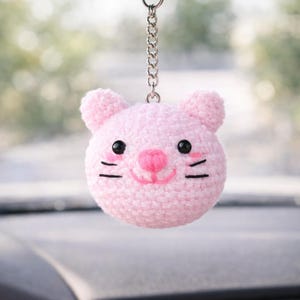 May include: A pink crocheted cat face car charm suspended from a silver chain. The cat features black eyes, whiskers, and a pink nose and mouth. The charm is hanging in front of a blurred background.