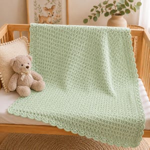 Könnte beinhalten: Eine hell salbeigrüne gehäkelte Babydecke mit strukturiertem Stichmuster und einem gewellten Rand. Die Decke liegt über dem Gitter eines Holzbettchens neben einem beigen Plüschbären, der eine cremefarbene Schleife um den Hals trägt.