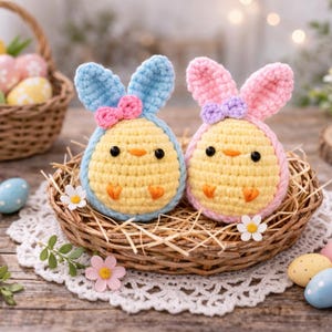 Könnte beinhalten: Zwei handgefertigte Häkel-Osterküken-Dekorationen mit blauen und rosa Hasenohren, in einem kleinen gewebten Nest. Die Küken sind gelb mit orangefarbenen Schnäbeln und schwarzen Augen. Ein Korb mit dekorierten Ostereiern ist im Hintergrund.