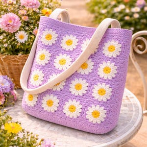 Könnte beinhalten: Eine lavendelfarbene Häkeltasche mit weißen und gelben Gänseblümchen-Applikationen. Die Tasche hat cremefarbene Träger und ist vor einem Hintergrund aus Blumen und Grün platziert, was auf ein Frühlings- oder Sommerthema hindeutet.
